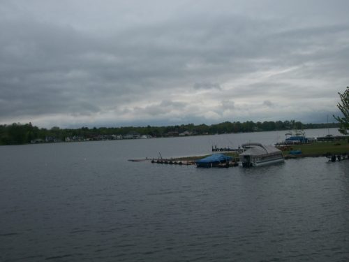 A calm lake scene with cloudy skies, boats docked, and green trees lining the shore.