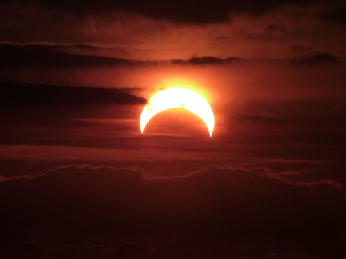 A partial solar eclipse with a crescent sun peeking through dark clouds at sunset.