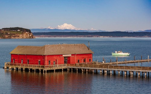 A red building on a pier with a boat nearby, set against a calm water backdrop and snow-capped mountains in the distance.