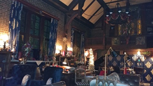 Cozy, dimly lit room with vintage furniture, decorated for the holidays, featuring a piano and large windows.