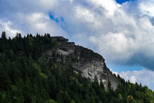 A rocky mountain peak rises above a dense forest under a partly cloudy sky.