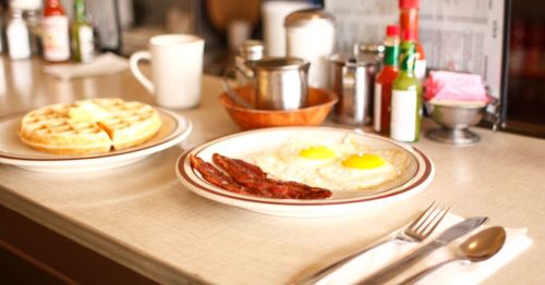 a plate of fried eggs and bacon next to a plate of waffles at a diner restaurant, with menus, hot sauce, and other condiments in the background