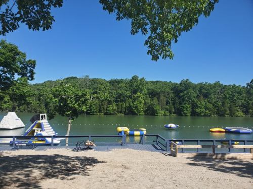 A serene lake scene with inflatable water toys, a climbing structure, and lush green trees under a clear blue sky.