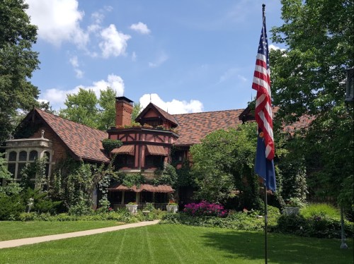 A large, historic mansion surrounded by greenery and flowers, with an American flag in the foreground.