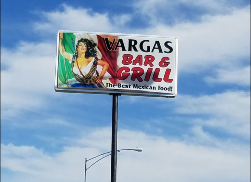 Sign for Vargas Bar & Grill featuring a woman holding a flag, promoting "The Best Mexican Food!" against a blue sky.