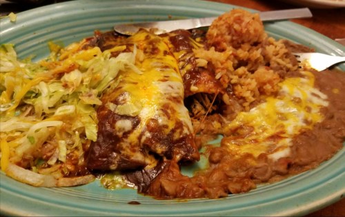 A plate of enchiladas topped with cheese, served with rice, refried beans, and a side of shredded lettuce.