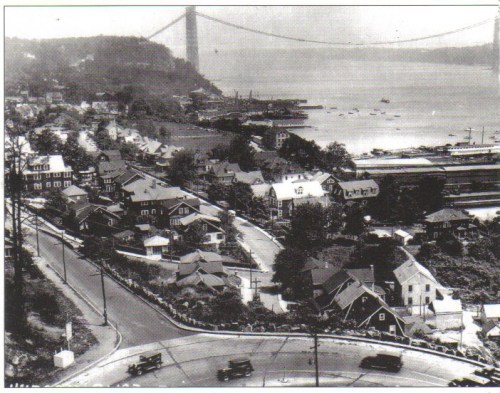 Aerial view of a waterfront neighborhood with houses, roads, and a bridge in the background, circa early 20th century.