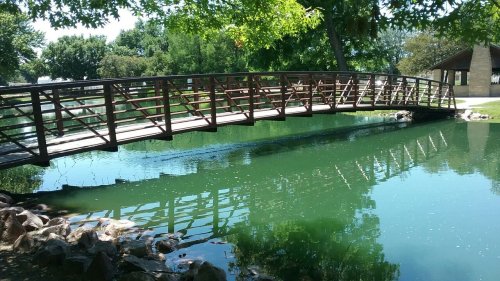 A wooden bridge arches over a calm green pond, surrounded by trees and rocks.