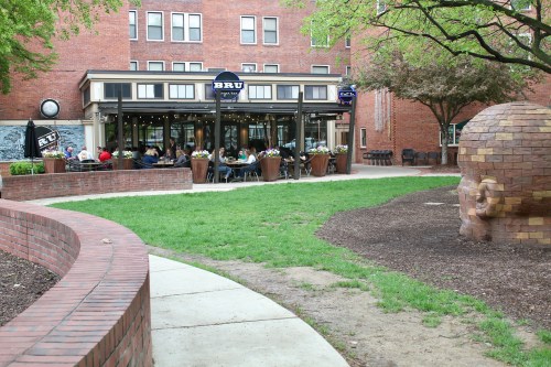 A café with outdoor seating surrounded by greenery and a brick pathway, featuring a large stone sculpture nearby.