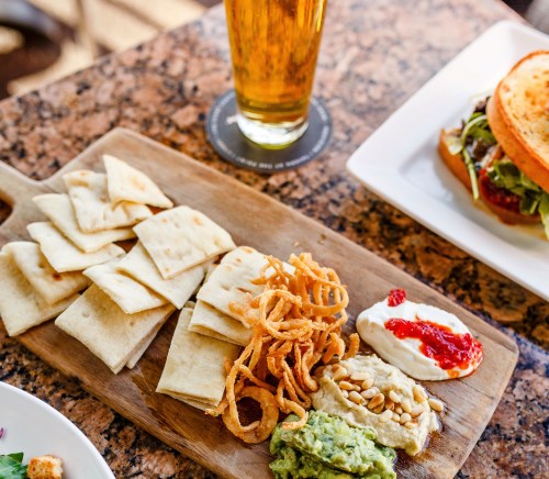 A wooden platter with pita chips, dips, and crispy onions, alongside a glass of beer and a sandwich on a table.