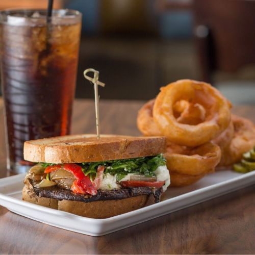 A hearty sandwich with grilled vegetables on toasted bread, served with onion rings and a glass of iced drink.