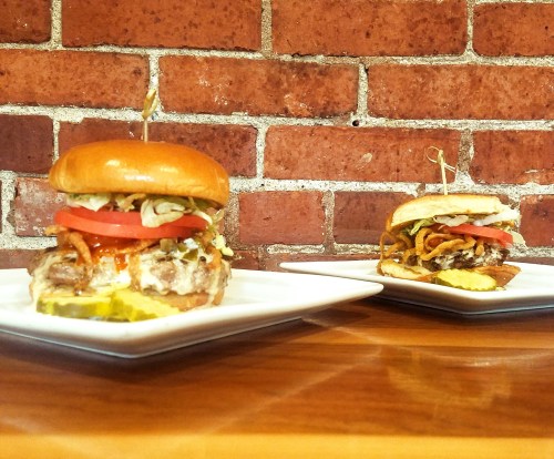 Two delicious burgers on white plates, topped with lettuce, tomato, pickles, and crispy onions, against a brick wall.