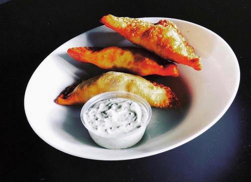 Three golden-brown pastries on a white plate, served with a small cup of creamy dipping sauce.