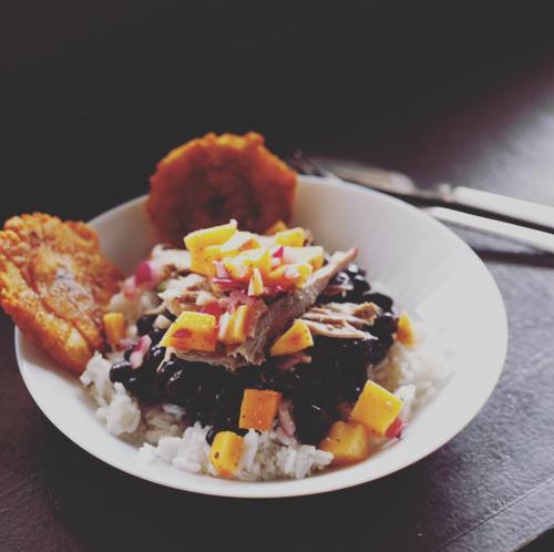 A bowl of rice topped with black beans, shredded meat, mango, and crispy plantain slices.