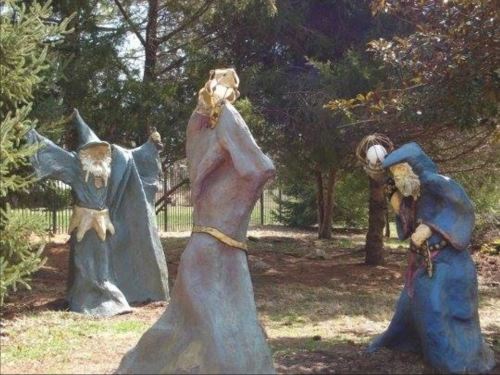 Three whimsical, colorful wizard sculptures in a wooded outdoor setting, each with unique poses and expressions.