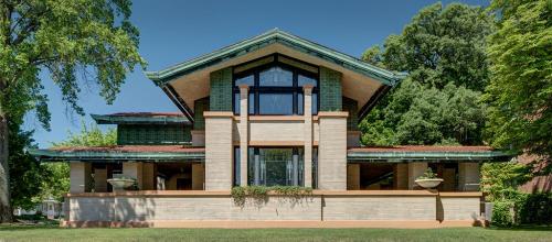 A large, modernist house with a distinctive roof, surrounded by trees and a green lawn under a clear blue sky.