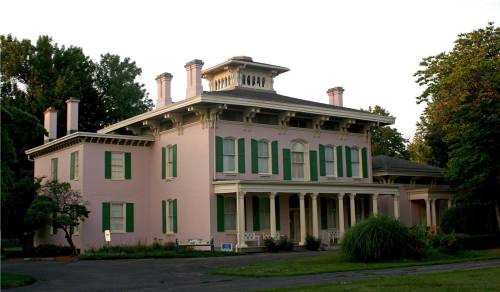 A large pink historic mansion with green shutters, surrounded by trees and a well-maintained garden.