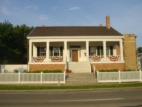 A historic white house with a porch, decorated with red, white, and blue bunting, surrounded by a white picket fence.