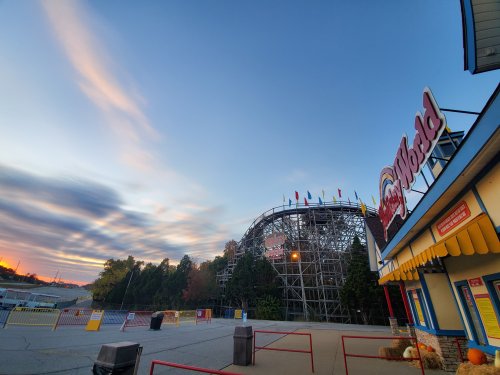 A colorful amusement park entrance with a wooden roller coaster in the background under a vibrant sky at dusk.