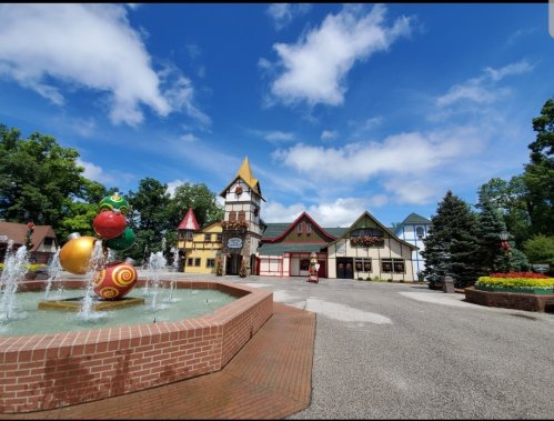 Colorful buildings with a fountain in the foreground, set against a bright blue sky and lush green trees.
