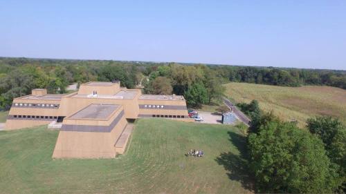 Aerial view of a modern building surrounded by trees and open fields, with a small group of people in the foreground.