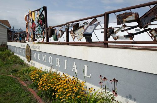 A colorful memorial with various objects displayed on a fence, surrounded by flowers and a cloudy sky.