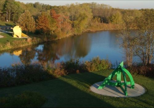 A vibrant green sculpture stands by a calm lake, surrounded by trees in autumn colors under a clear sky.