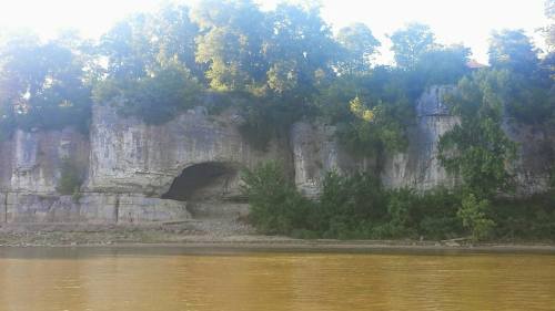 A rocky cliff with a large cave opening, surrounded by trees, beside a calm river.