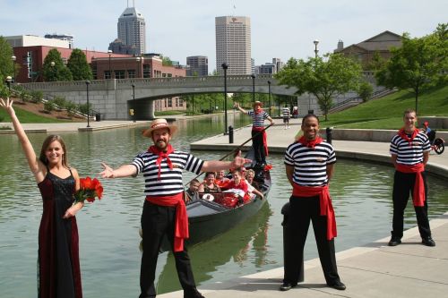 A group of performers in striped shirts and red scarves pose by a river, with a boat of people in the background.