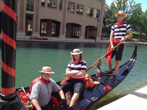 Three men in striped shirts and hats pose on a gondola by a canal, surrounded by greenery and flowers.