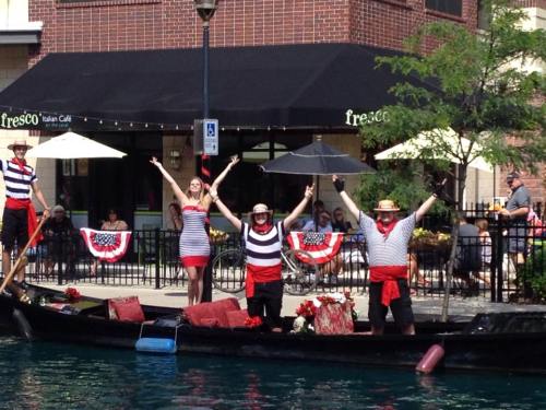 A group of performers in striped outfits celebrate on a gondola, with a café and patrons in the background.