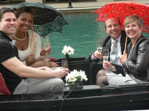 A group of four people enjoying a boat ride, holding champagne glasses and umbrellas, with flowers in the background.