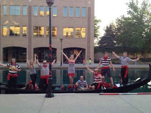 A group of people in striped shirts and red accents joyfully pose on a gondola by a canal, with buildings in the background.