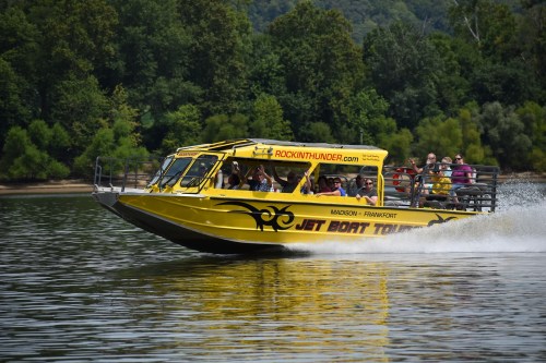 A yellow jet boat speeds across a river, with passengers enjoying the ride against a backdrop of green trees.