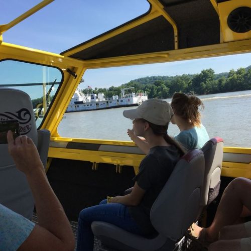 Two girls sit inside a yellow boat, looking out at a passing vessel on a river, surrounded by green hills.