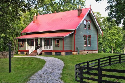 A charming two-story house with a red roof, surrounded by greenery and a gravel path leading to a black fence.