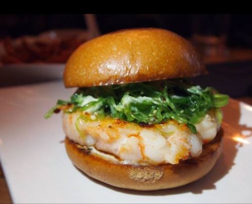 A close-up of a shrimp burger with a soft bun, topped with green seaweed salad on a white plate.