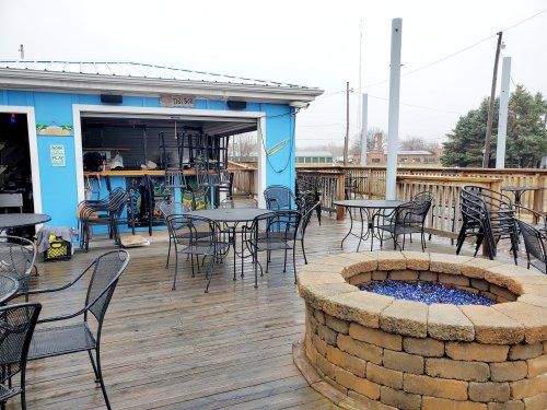 Outdoor seating area with black chairs and tables, a stone fire pit, and a blue bar building in the background.