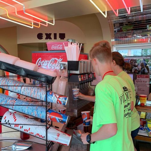 Two young people in bright green shirts fill cups at a soda fountain in a colorful diner setting.