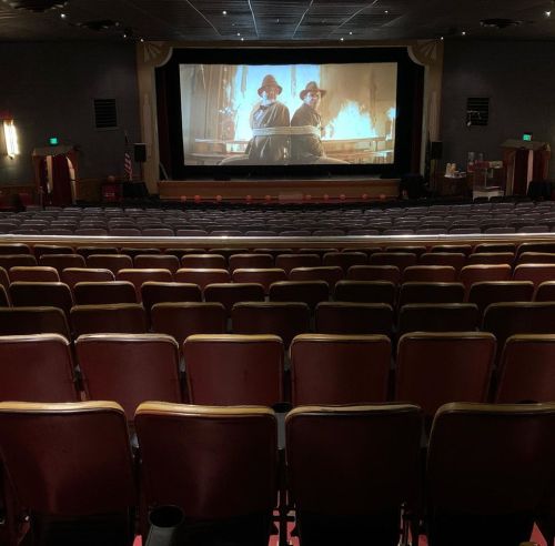 A nearly empty movie theater with red seats, featuring a large screen showing two characters in a dramatic scene.