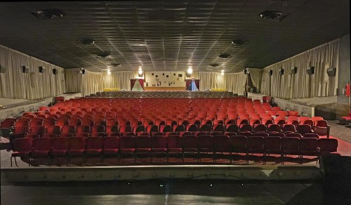 A dimly lit theater with rows of red seats facing a stage, surrounded by white curtains and soft lighting.