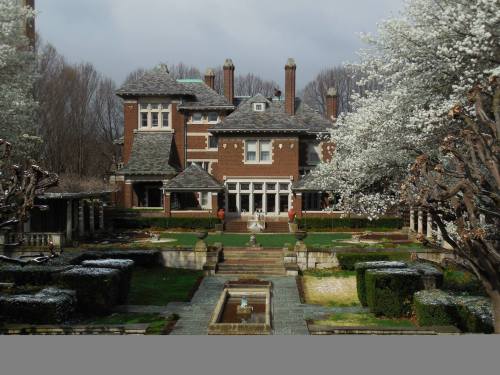 A grand brick mansion surrounded by manicured gardens and blooming trees under a cloudy sky.