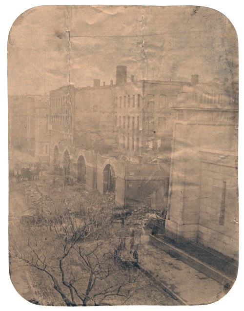 Faded historical photo of a street scene with buildings and trees, showing signs of age and wear.
