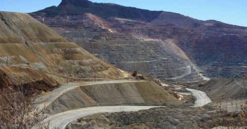 A vast open-pit mine with layered earth and winding roads, set against a mountainous backdrop under a clear blue sky.