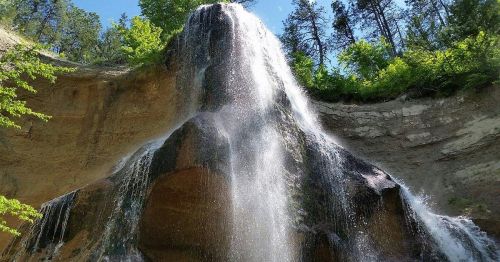 A cascading waterfall flows down a rocky cliff surrounded by lush green trees under a clear blue sky.