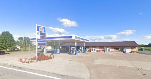 A gas station with a convenience store, featuring fuel pumps and a price sign under a clear blue sky.