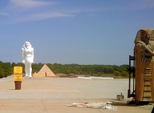 A large white statue of a pharaoh stands beside a pyramid and a smiling sign in a wide, open area.