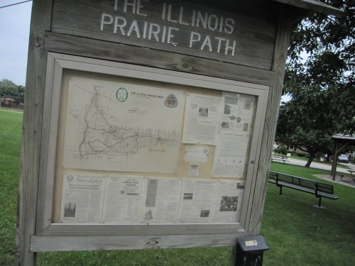 Information board displaying a map and newsletters about the Illinois Prairie Path in a grassy area.