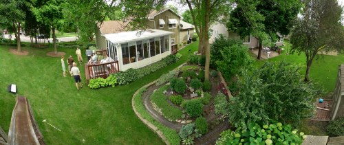 A panoramic view of a lush backyard with a garden, a deck, and people socializing near a house.