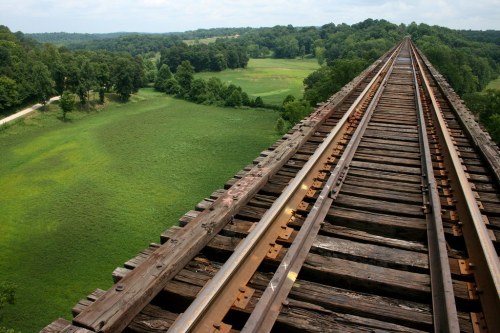 Aerial view of a wooden railway bridge stretching over lush green fields and trees under a cloudy sky.